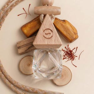 May include: A clear glass perfume bottle with a wooden triangle top and wooden accents. The bottle is surrounded by wood slices, saffron threads, and a rope with gold accents. The wooden triangle has a circular logo.