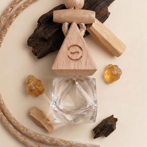 May include: A wooden car air freshener with a triangular design and a clear glass bottle. The freshener is suspended by a beige rope with gold accents. The image also includes pieces of wood and amber-colored stones.