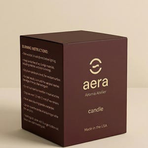 May include: A dark maroon square candle box featuring the gold Aera Aroma Atelier logo and the word candle. The side panel displays burning instructions, including specific measurements like 1/4 inch and 1/2 inch, and states Made in the USA.