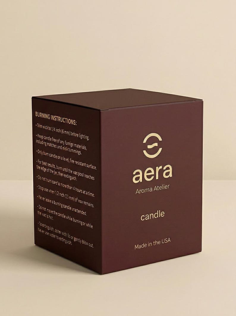 May include: A dark maroon square candle box featuring the gold Aera Aroma Atelier logo and the word candle. The side panel displays burning instructions, including specific measurements like 1/4 inch and 1/2 inch, and states Made in the USA.