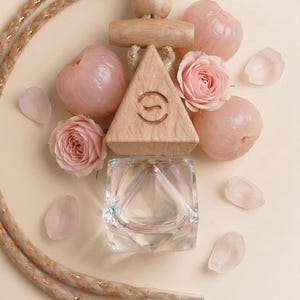 May include: A wooden triangle pendant with a carved design, a clear glass container, and a decorative rope. The image also features pink roses, lychees, and petals, all arranged on a cream-coloured surface.
