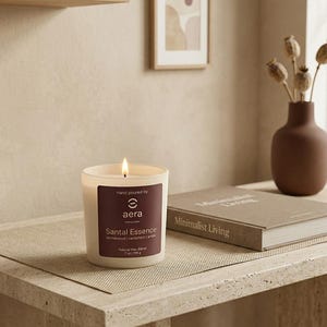 May include: A lit candle in a white frosted glass container with a dark brown label. The label reads Hand poured by aera Santal Essence, sandalwood | cardamom | amber, Natural Wax Blend, 7 oz | 198 g. The candle has a single cotton wick.