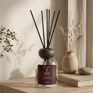 Peut inclure: Un diffuseur de parfum en verre avec un bouchon en bois fonc&eacute; et des b&acirc;tonnets noirs. L'&eacute;tiquette indique "aera Aroma Atelier Ruby Cherry". Un petit vase avec des plantes s&eacute;ch&eacute;es et un livre sont &eacute;galement sur la surface en bois.