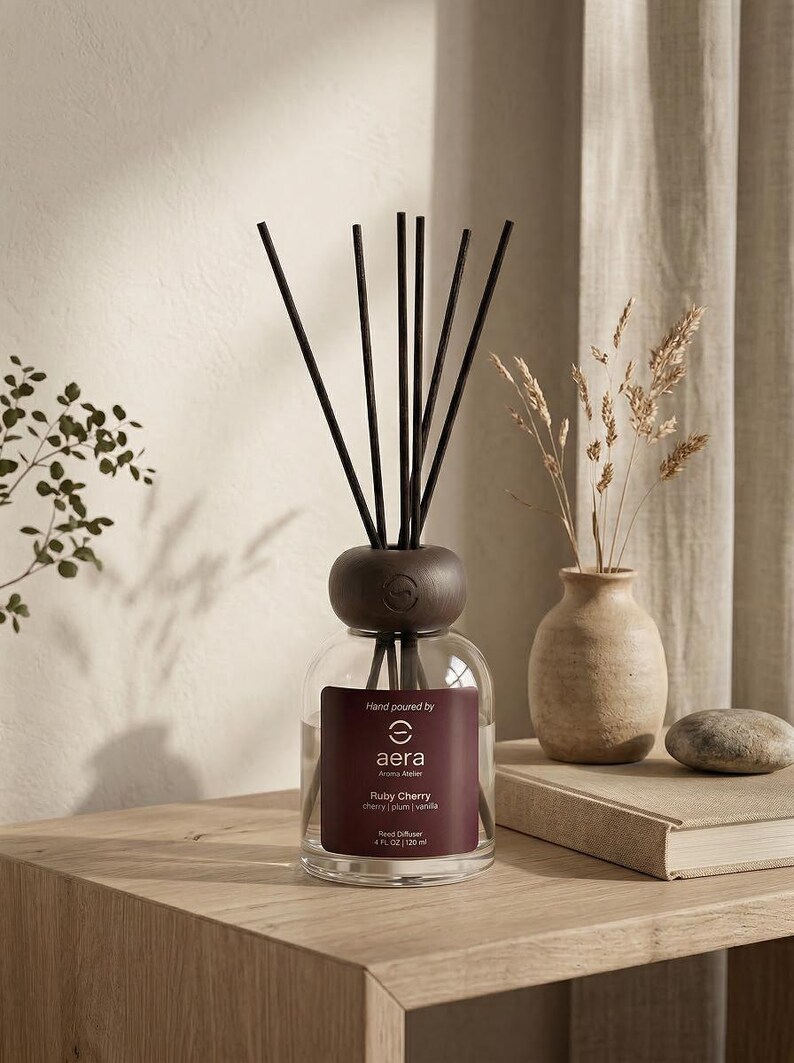 Peut inclure: Un diffuseur de parfum en verre avec un bouchon en bois fonc&eacute; et des b&acirc;tonnets noirs. L'&eacute;tiquette indique "aera Aroma Atelier Ruby Cherry". Un petit vase avec des plantes s&eacute;ch&eacute;es et un livre sont &eacute;galement sur la surface en bois.