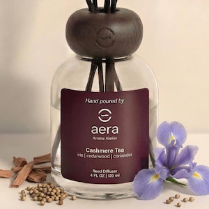 May include: A clear glass reed diffuser bottle with a dark brown cap and reeds. The label reads "Cashmere Tea" and "Aera Aroma Atelier." A purple iris flower and wood pieces are in the foreground. The bottle contains 4 fl oz / 120 ml.