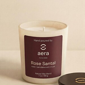 May include: A white frosted glass candle with a dark maroon label reading Hand poured by aera Aroma Atelier Rose Santal rose | sandalwood | musk Natural Wax Blend 7 oz | 198 g. A matching dark maroon lid with a gold logo sits beside the candle.