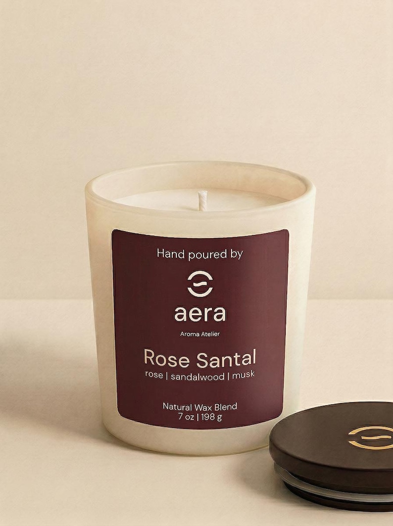 May include: A white frosted glass candle with a dark maroon label reading Hand poured by aera Aroma Atelier Rose Santal rose | sandalwood | musk Natural Wax Blend 7 oz | 198 g. A matching dark maroon lid with a gold logo sits beside the candle.