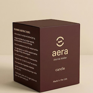 May include: A dark maroon square candle box featuring the gold Aera Aroma Atelier logo and the word candle. The side panel displays burning instructions, including specific measurements like 1/4 inch and 1/2 inch, and states Made in the USA.