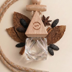 May include: A clear glass perfume bottle with a faceted geometric base and a light wood triangular cap featuring an engraved letter S. The bottle hangs from a braided beige cord with gold metallic threads, surrounded by dried tobacco leaves.