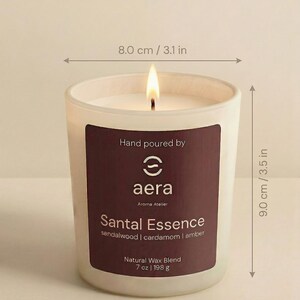 May include: A lit candle in a frosted glass jar with a dark label reading Hand poured by aera Aroma Atelier Santal Essence sandalwood | cardamom | amber Natural Wax Blend 7 oz. The jar measures 3.1 inches wide at the top, 2.9 inches at the base, and 3.5 inches tall.