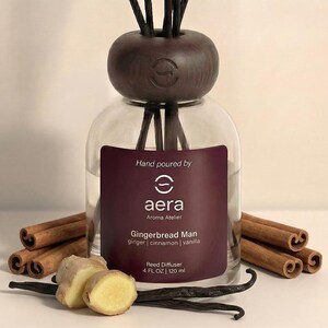 May include: A clear glass reed diffuser with a dark brown wooden cap and reeds. The label reads "Gingerbread Man" with ginger, cinnamon, and vanilla scents. The bottle is surrounded by cinnamon sticks, ginger slices, and vanilla beans. The diffuser contains 4 fl oz / 120 ml.