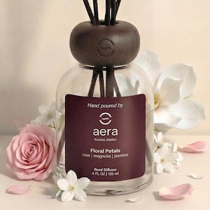 May include: A clear glass reed diffuser with a dark brown wooden cap and reeds. The label reads "Floral Petals" and lists rose, magnolia, and jasmine. The bottle is surrounded by pink rose and white magnolia flowers.