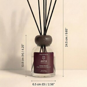 K&ouml;nnte beinhalten: Ein klarer Glas-Reed-Diffusor mit einem dunkelbraunen Holzaufsatz und Schilf. Auf dem Etikett steht "aera Aroma Atelier Amber Smoke". Der Diffusor ist 10,8 cm hoch und 6,5 cm im Durchmesser, mit einem Nettoinhalt von 120 ml.