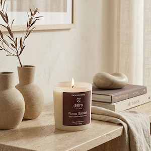 May include: A lit candle in a clear glass jar with a dark brown label reading Hand poured by aera Rose Santal rose | sandalwood | musk Natural Wax Blend 7oz | 198g. The candle is placed on a textured stone surface next to two beige vases.