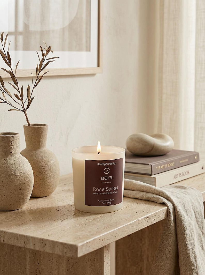 May include: A lit candle in a clear glass jar with a dark brown label reading Hand poured by aera Rose Santal rose | sandalwood | musk Natural Wax Blend 7oz | 198g. The candle is placed on a textured stone surface next to two beige vases.