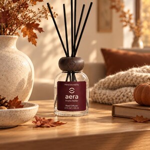 Puede incluir: Un difusor de varillas de vidrio transparente con tapa de madera oscura y varillas negras. La etiqueta es de color rojo oscuro con texto blanco que dice Hand poured by aera Aroma Atelier Reed Diffuser 4 FL OZ | 120 ml. Contiene líquido.
