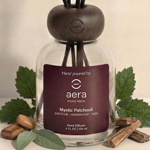May include: A clear glass reed diffuser with a dark brown wooden top and reeds. The label reads "Mystic Patchouli" with patchouli, sandalwood, and sage scents. The bottle contains 4 fl oz, or 120 ml. Green leaves and wood pieces surround the diffuser.
