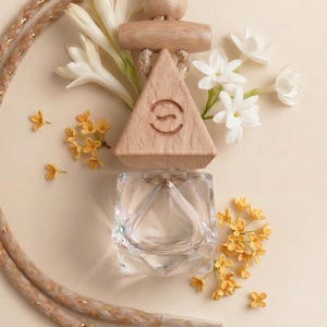 May include: A clear glass perfume bottle with a wooden top, featuring a triangle shape with a curved design. The bottle is surrounded by white and yellow flowers, and a decorative rope. The background is a light beige colour.