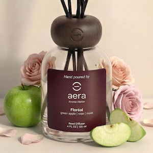 May include: A clear glass reed diffuser with a dark brown wooden top and reeds. The label reads "aera Aroma Atelier Floréal green apple | rose | musk." A green apple and rose petals are in the foreground. The diffuser is 120 ml.