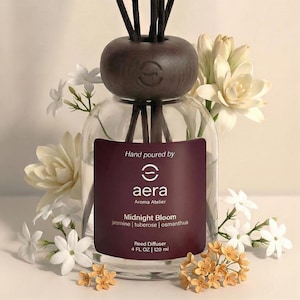 May include: A clear glass reed diffuser bottle with a dark brown wooden top and reeds. The label reads "Midnight Bloom" and "jasmine | tuberose | osmanthus". The bottle contains 120 ml of liquid and is surrounded by white and yellow flowers.