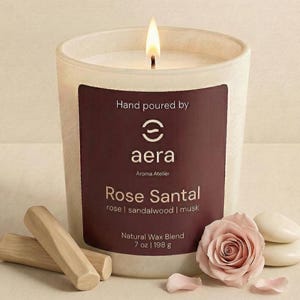 May include: A lit candle in a white frosted glass vessel with a dark maroon label reading Hand poured by aera Aroma Atelier Rose Santal rose | sandalwood | musk Natural Wax Blend 7 oz | 198 g, accompanied by two wooden sticks and a pink rose.