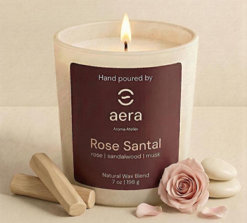 May include: A lit candle in a white frosted glass vessel with a dark maroon label reading Hand poured by aera Aroma Atelier Rose Santal rose | sandalwood | musk Natural Wax Blend 7 oz | 198 g, accompanied by two wooden sticks and a pink rose.
