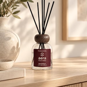 Puede incluir: Un difusor de varillas de vidrio transparente con un cuello de madera oscura y seis varillas negras. La etiqueta dice Hand poured by aera Aroma Atelier Reed Diffuser 4 FL OZ | 120 ml. El frasco contiene un líquido claro y una pieza de madera.