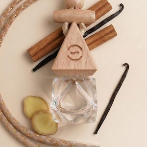 May include: An air freshener with a wooden triangle and a clear glass container. The wooden triangle has a logo. Cinnamon sticks, vanilla beans, and ginger slices are arranged around the air freshener, with a decorative rope.