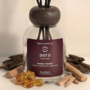 May include: A clear glass reed diffuser with a dark brown wooden top and reeds. The label reads "Amber Smoke" with amber, sandalwood, and agarwood scents. The bottle contains 4 fl oz (120 ml).