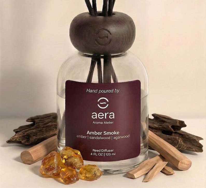 K&ouml;nnte beinhalten: Ein klarer Glas-Reed-Diffusor mit einem dunkelbraunen Holzaufsatz und Schilf. Das Etikett lautet "Amber Smoke" mit Amber-, Sandelholz- und Agarwood-D&uuml;ften. Die Flasche enth&auml;lt 120 ml.