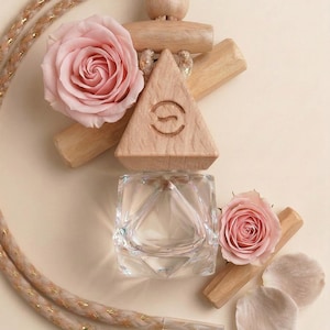 May include: An arrangement featuring a clear glass square-shaped container, a wooden triangle with a logo, and two pink roses. A braided rope with gold accents and wooden accents are also present. Rose petals complete the composition.