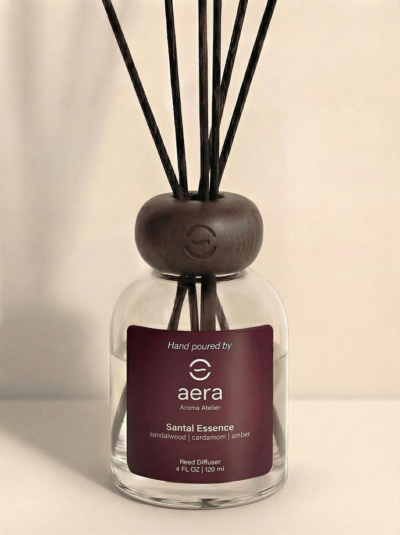 May include: A clear glass reed diffuser with a dark brown wooden top and reeds. The label reads "aera Aroma Atelier Santal Essence" with sandalwood, cardamom, and amber notes. The bottle contains 120 ml.