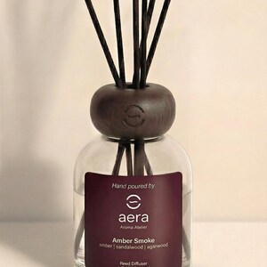 K&ouml;nnte beinhalten: Ein klarer Glas-Reed-Diffusor mit einem dunkelbraunen Holzaufsatz und Schilf. Das Etikett lautet "aera Aroma Atelier Amber Smoke" mit D&uuml;ften von Amber, Sandelholz und Adlerholz. Die Flasche enth&auml;lt 120 ml Duft.