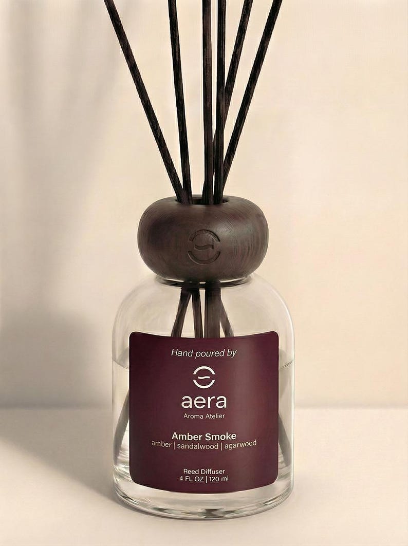K&ouml;nnte beinhalten: Ein klarer Glas-Reed-Diffusor mit einem dunkelbraunen Holzaufsatz und Schilf. Das Etikett lautet "aera Aroma Atelier Amber Smoke" mit D&uuml;ften von Amber, Sandelholz und Adlerholz. Die Flasche enth&auml;lt 120 ml Duft.