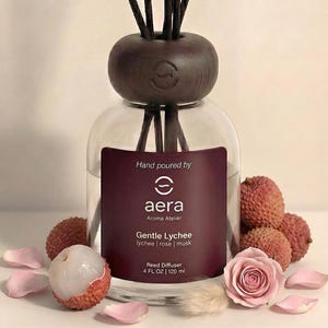 May include: A clear glass reed diffuser with a dark brown wooden top and reeds. The label reads "aera Aroma Atelier Gentle Lychee" and "120 ml". Lychee fruit, rose petals, and a pink rose are arranged around the diffuser.