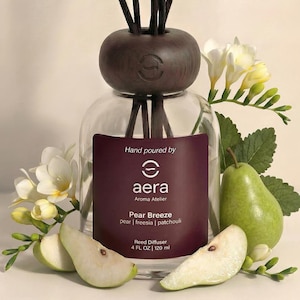 May include: A clear glass reed diffuser with a dark brown wooden top and reeds. The label reads "Pear Breeze" and "4 FL OZ | 120 ml". Sliced pears and white flowers are arranged around the diffuser.