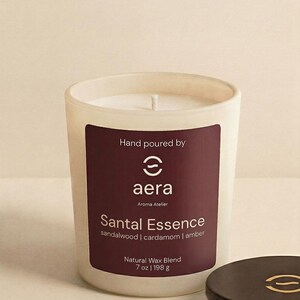May include: A white frosted glass candle with a dark maroon label featuring the text Hand poured by aera Aroma Atelier Santal Essence sandalwood | cardamom | amber Natural Wax Blend 7 oz | 198 g and a separate dark brown circular lid.