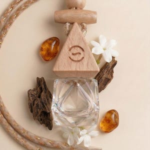 May include: A wooden car air freshener with a geometric design, a clear glass bottle, amber stones, white flowers, and wood pieces. The air freshener is suspended by a beige and gold rope. The design includes a triangle with a logo.