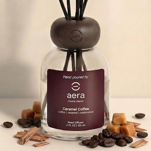 May include: A clear glass reed diffuser with a dark brown wooden cap. The label reads "Caramel Coffee" with the brand name "aera". The diffuser contains black reeds and is surrounded by coffee beans, caramel candies, and wood chips. The bottle is 4 fl oz / 120 ml.