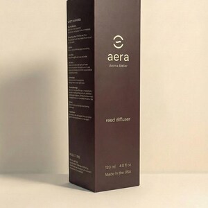 May include: A dark brown rectangular box for a reed diffuser, branded with "aera" and "Aroma Atelier" in white. The box states a volume of 120 ml (4.0 fl oz) and "Made in the USA". Safety warnings are printed on the side of the box.