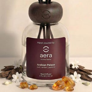 May include: A clear glass reed diffuser with a dark brown wooden cap and reeds. The label reads "aera Arabian Palace oud | amber | jasmine." The diffuser is surrounded by white flowers, amber stones, and wood pieces. The bottle contains 4 fl oz / 120 ml.