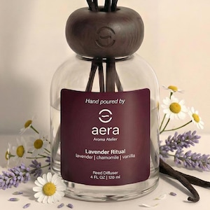 May include: A clear glass reed diffuser bottle with a dark brown cap and reeds. The label reads "aera Aroma Atelier Lavender Ritual" with 4 fl oz / 120 ml. The bottle is surrounded by lavender, chamomile, and vanilla.