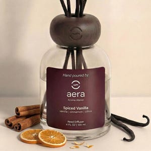 May include: A clear glass reed diffuser bottle with a dark wood cap and black reeds. The label reads Hand poured by aera Aroma Atelier Spiced Vanilla vanilla | cinnamon | citrus Reed Diffuser 4 FL OZ | 120 ml. Cinnamon sticks and dried orange slices.