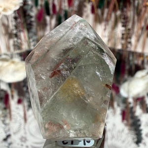 May include: A clear quartz crystal with a faceted, pointed top. The crystal has internal inclusions of red and green, and a slight yellow hue. The crystal is displayed on a reflective surface.