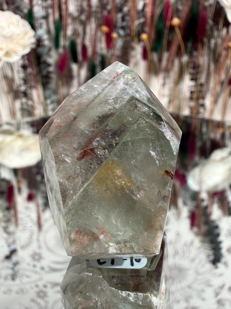 May include: A clear quartz crystal with a faceted, pointed top. The crystal has internal inclusions of red and green, and a slight yellow hue. The crystal is displayed on a reflective surface.