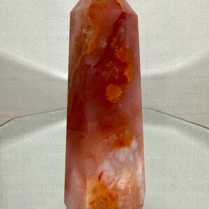 May include: A polished crystal tower with a hexagonal base and a pointed top. The stone features a translucent appearance with swirling patterns of deep orange, burnt red, and milky white inclusions throughout the entire vertical structure.