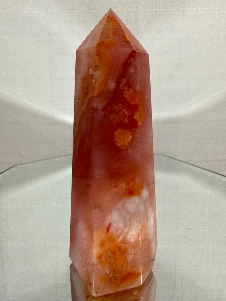 May include: A polished crystal tower with a hexagonal base and a pointed top. The stone features a translucent appearance with swirling patterns of deep orange, burnt red, and milky white inclusions throughout the entire vertical structure.