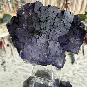 Puede incluir: Un gran grupo de cristales de fluorita de color p&uacute;rpura oscuro con una estructura c&uacute;bica. El cristal se exhibe sobre un soporte acr&iacute;lico transparente, mostrando su forma natural y variaciones de color. La piedra tiene una superficie texturizada.