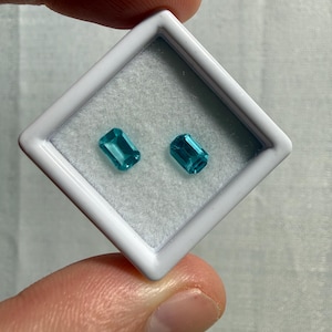 May include: Two emerald-cut, vibrant teal-blue gemstones are displayed inside a square white plastic display case with a soft, light gray velvet-like insert. The stones are positioned side-by-side, showcasing their faceted surfaces and clarity.