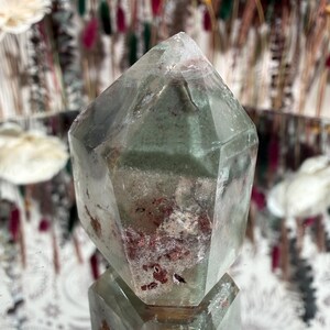 May include: A clear quartz crystal with green and red inclusions. The crystal is faceted with a pointed top and a flat base. The crystal is sitting on a reflective surface. The background is blurred with floral elements.
